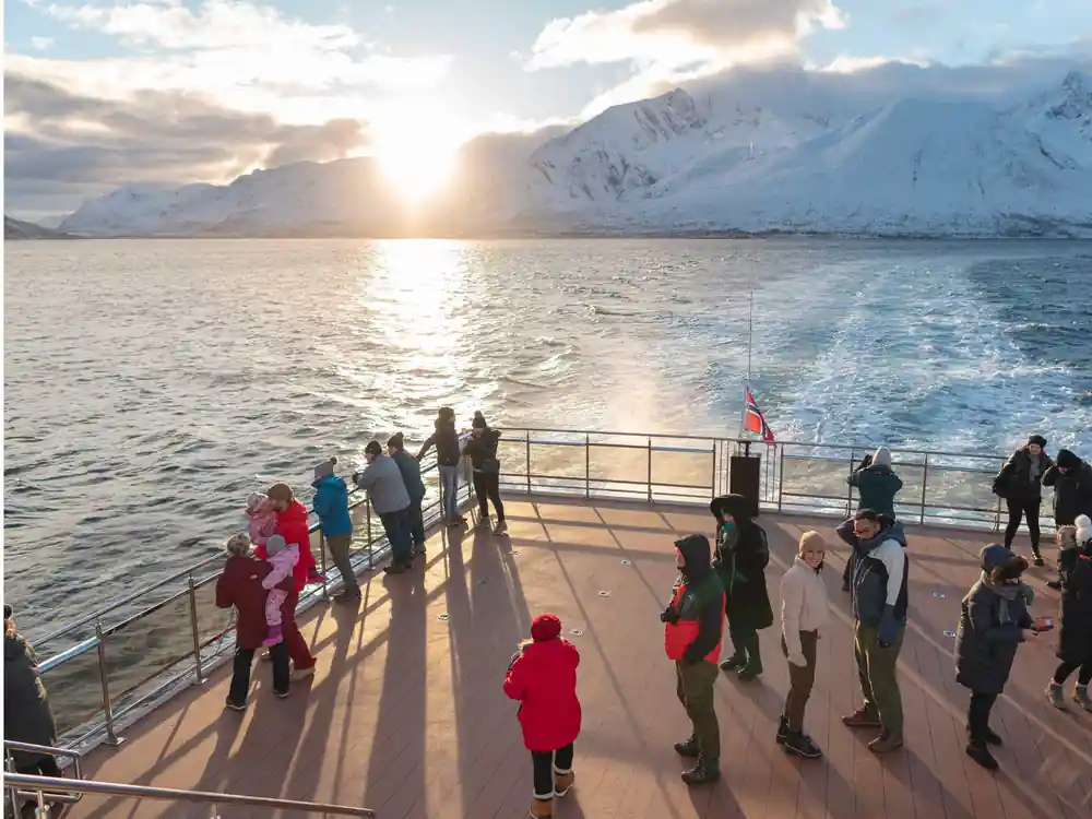 Passengers on ship deck viewing snowy mountains at sunset